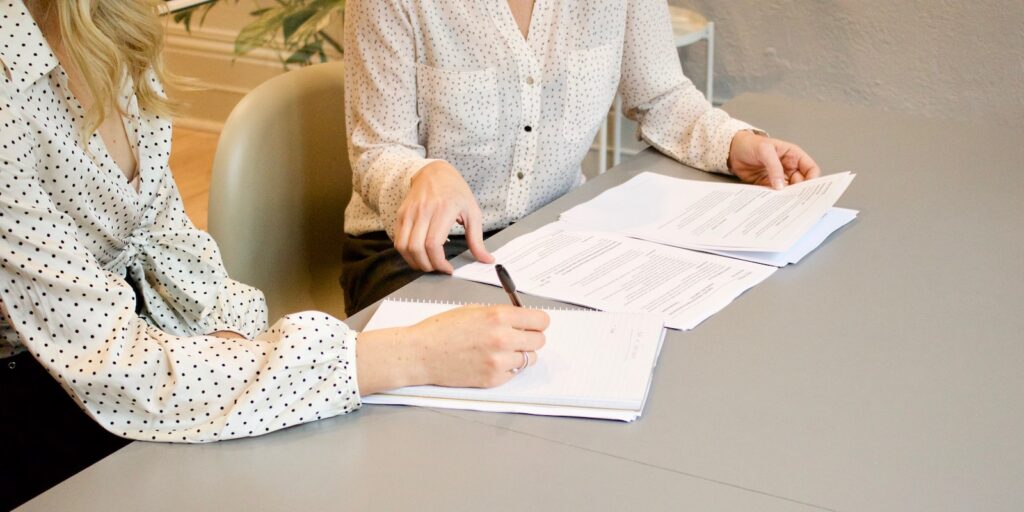 negotiation, woman signing on white printer paper beside woman about to touch the documents